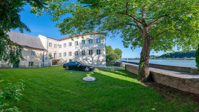 Au&szlig;enansicht eines alten Postamts mit Baum und Auto im Vordergrund, Donau im Hintergrund.