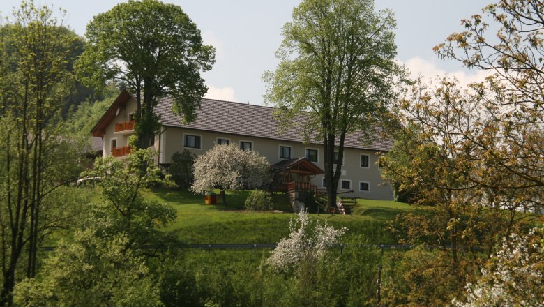 Karhof - Urlaub am Bauernhof, © Archiv Betrieb Ein Bauernhof auf einem grünen Hügel, umgeben von Bäumen und blühenden Sträuchern.