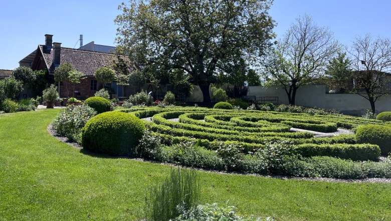 Ein gepflegter Garten mit einem Labyrinth aus Hecken, einem großen Baum und einem kleinen Gebäude im Hintergrund bei sonnigem Wetter.