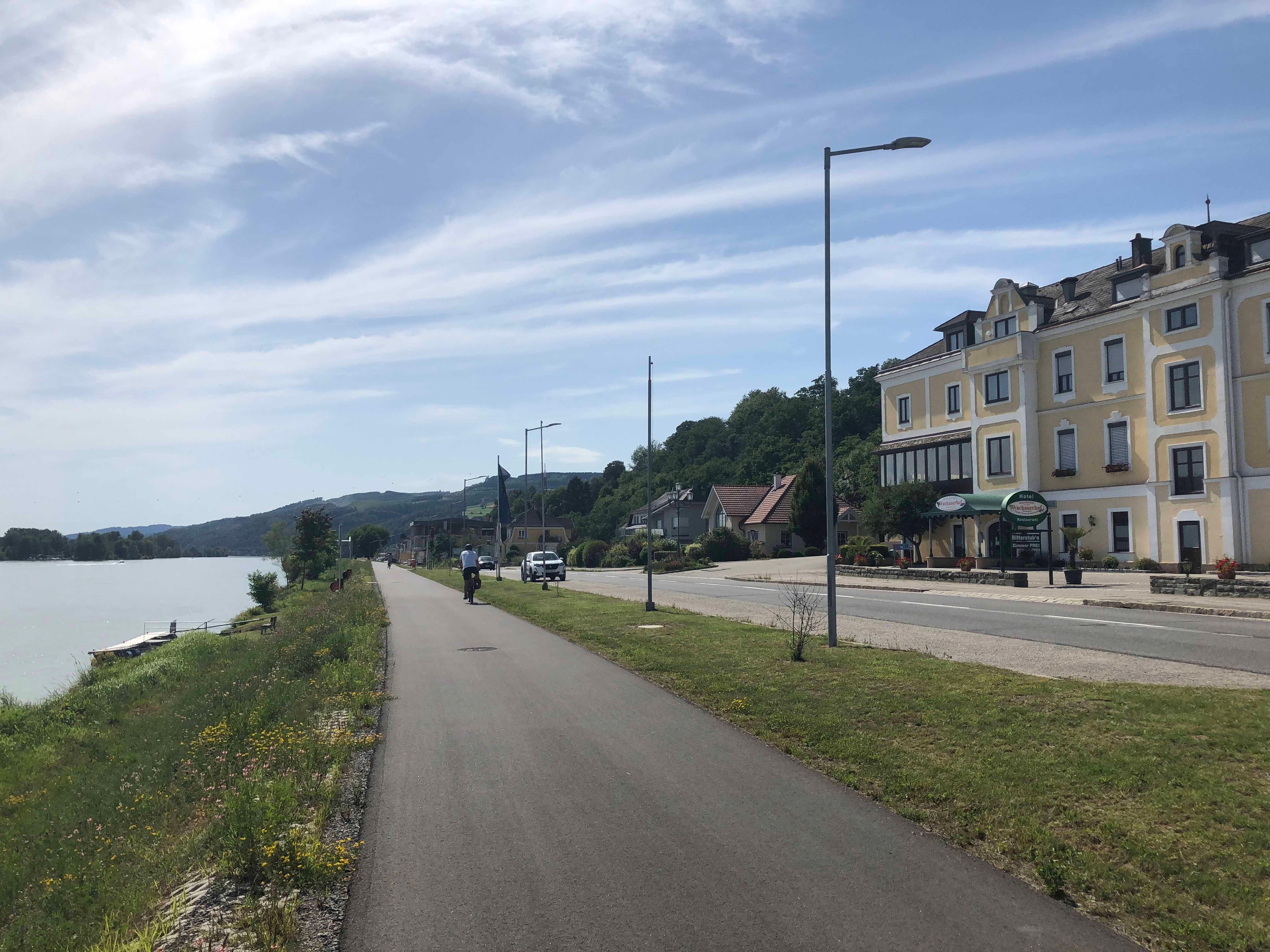 Straße am Flussufer mit gelbem Gebäude und Radfahrer.