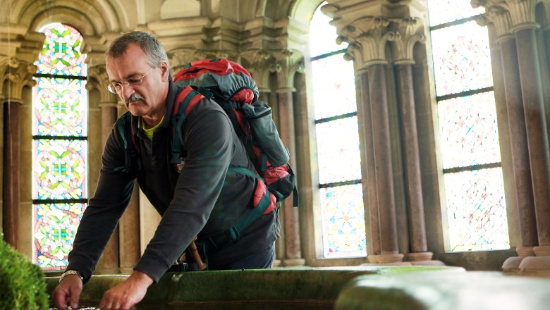 Ein Mann mit Rucksack beugt sich über ein Wasserbecken in einem historischen Gebäude mit bunten Glasfenstern.