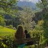 Eine Frau sitzt mit einem Hund auf einer Bank und blickt auf eine gr&uuml;ne Landschaft mit bl&uuml;henden B&auml;umen und Bergen im Hintergrund.