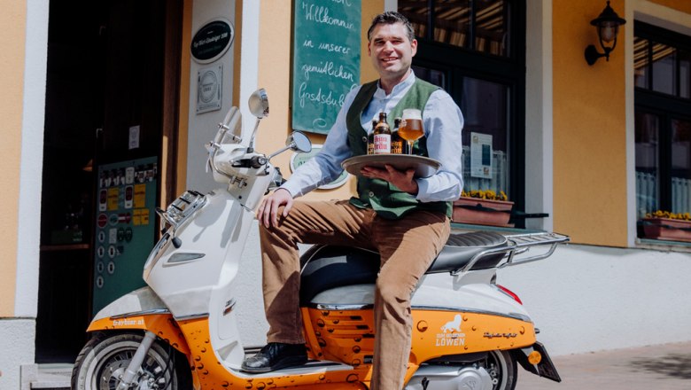 Ein Mann sitzt auf einem Roller vor einem Gebäude und hält ein Tablett mit Bierflaschen und einem Glas.