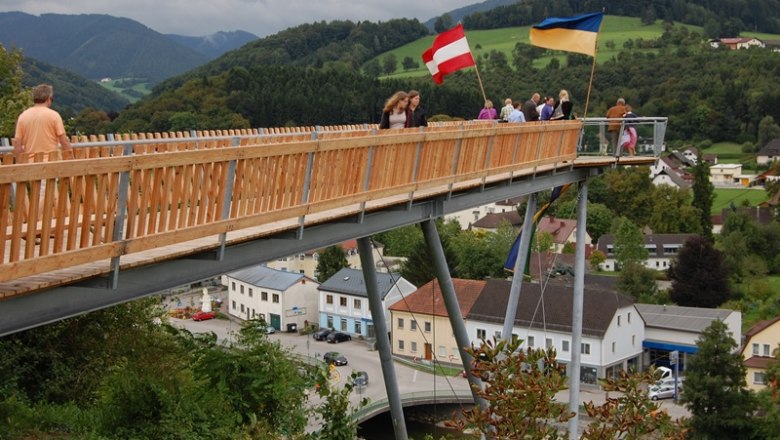 Menschen auf einer Holzplattform mit &ouml;sterreichischer und ukrainischer Flagge, im Hintergrund eine l&auml;ndliche Landschaft.