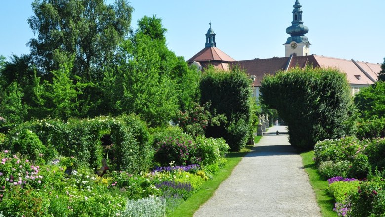 Ein gepflegter Gartenweg mit Blumen und Bäumen, im Hintergrund ein historisches Gebäude mit Türmen.