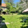 Ein gr&uuml;ner Garten mit bl&uuml;henden B&auml;umen, einem Sonnenschirm und einem alten Geb&auml;ude im Hintergrund.