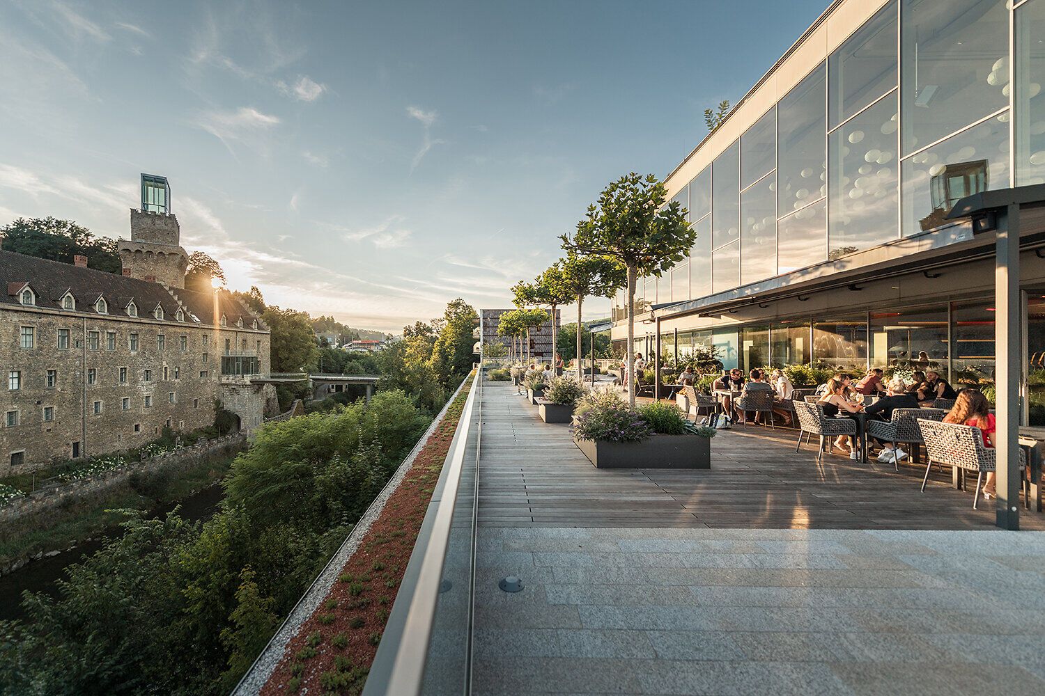 Moderne Restaurant-Dachterrasse mit Gästen bei Abendsonne. Im Hintergrund kontrastiert ein historisches Schlossgebäude mit dem modernen Glasbau des Vordergrunds.