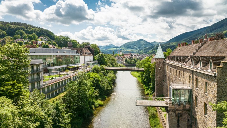 Panorama einer Flusslandschaft mit Schloss und moderner Architektur, umgeben von gr&uuml;nen H&uuml;geln.