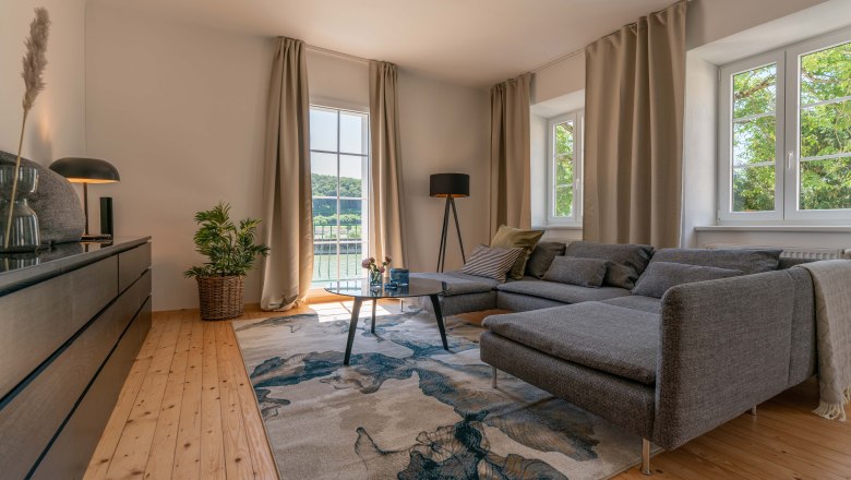 Modernes Wohnzimmer mit grauem Sofa, Holzboden und gro&szlig;em Fenster mit Blick ins Gr&uuml;ne.